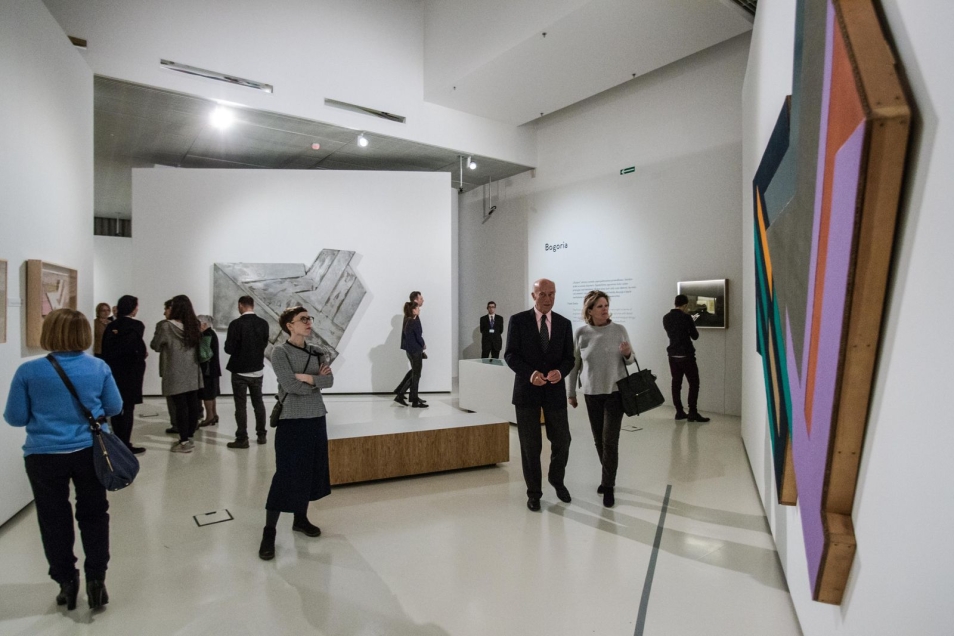Sala w muzeum. Wystawa „Frank Stella i synagogi dawnej Polski”. Na ścianach wiszą kolorowe obrazy. Oglądają je zebrani w sali ludzie.    