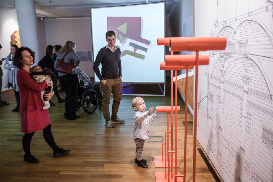 Sala w muzeum. Rodzina z dwójką małych dzieci ogląda eksponaty na wystawie „Frank Stella i synagogi dawnej Polski”. Na ścianie szkic przedstawiający synagogę. W tle duży, kolorowy obraz.  