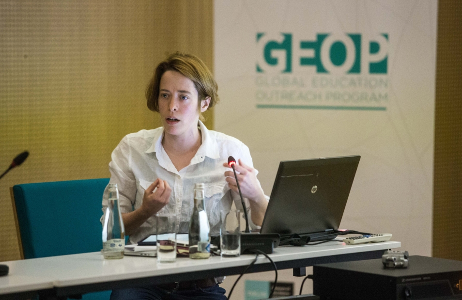 A woman in a white shirt is sitting at a table with an open laptop in front of her. She is speaking to people outside the frame of the photograph, with an advertising banner bearing the inscription GEOP in the background.