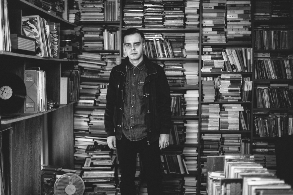 Daniel Drumz. Młody mężczyzna stoi na tle regałów z książkami.
