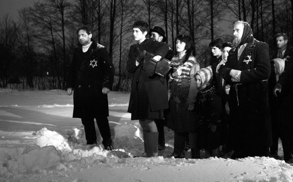 Czarno-białe zdjęcie. Grupa Żydów z gwiazdami Dawida wyhaftowanymi na płaszczach stoi na śniegu. 
