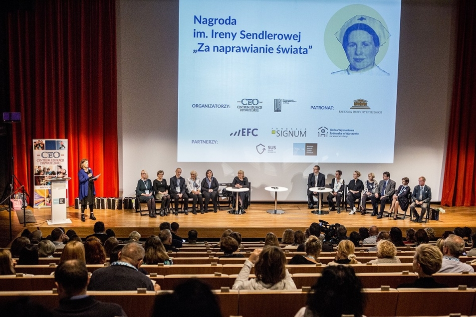Nagrody im. Ireny Sendlerowej "Za naprawianie świata", Muzeum POLIN, Widok sali wykładowej z perspektywy widowni. Na scenie siedzą paneliści, w dwóch grupach - po prawej i lewej. Nad ich głowami ekran, na którym wyświetlony jest slajd wykładu