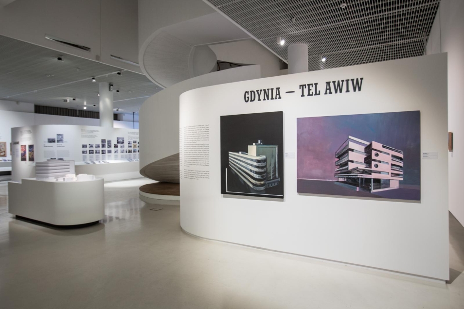 Poranne ptaszki - oprowadzanie po wystawie "Gdynia - Tel Awiw" dla osób ze spektrum autyzmu w Muzeum POLIN. Na zdjęciu przestrzeń wystawy, podzielona białymi ścianami działowymi, na pierwszym planie ściana z tytułem wystawy i dwoma obrazami w kolorystyce czarnej, fioletowej, przedstawiające budynki modernistyczne