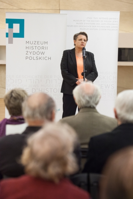 Kobieta stoi przy mikrofonie podczas wernisażu o Sprawiedliwych.