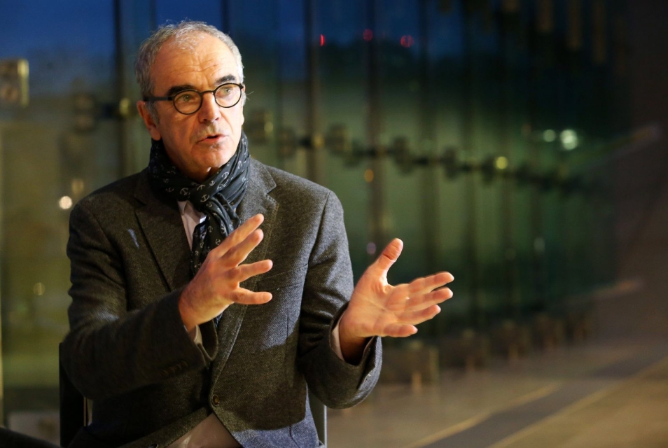 Sławomir Idziak - gestykuluje w dialogu z rozmówcą. W tle szklana ściana Muzeum POLIN, za oknem widać noc.