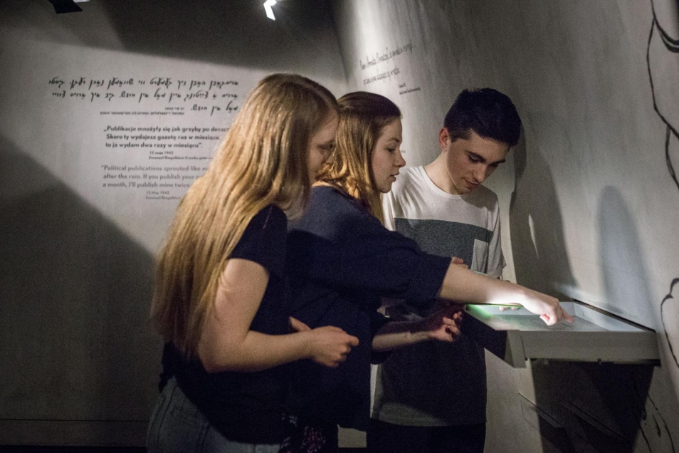 Dwie dziewczyny i chłopiec stoją przed gablotką w przestrzeni wystawy stałej Muzeum POLIN. W pomieszczeniu jest ciemno.