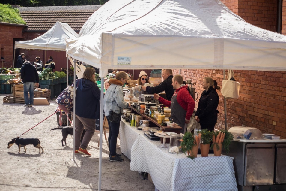 Targ w Fortecy Kręglickich w ramach TISZ Festiwalu Muzeum POLIN, Na zdjęciu: w otwartej przestrzeni, pod ścianami z czerwonych cegieł, rozstawione są stołychronione parasolami. Na stołach porozkładane produkty żywnościowe i warzywa oraz owoce. Wokół straganów ludzie - sprzedawcy i kupujący