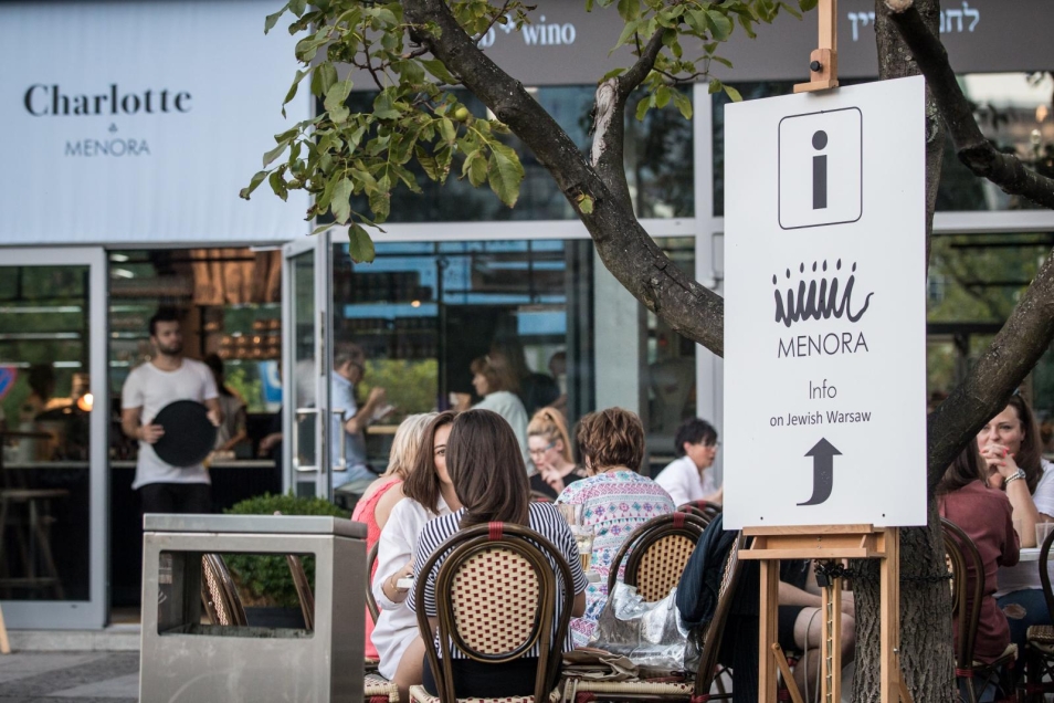 TISZ Festiwal: śniadanie po warszawsku. Na zdjęciu scena przed restauracją Charlotte Menora. Obok białej tabliczki z logo restauracji widać stoliki, przy których, przy śniadaniu, siedzą kobiety i mężczyźni. W tle, w drzwiach do restauracji, widać kelnera w białym podkoszulku. Trzyma czarną okrągłą tacę.