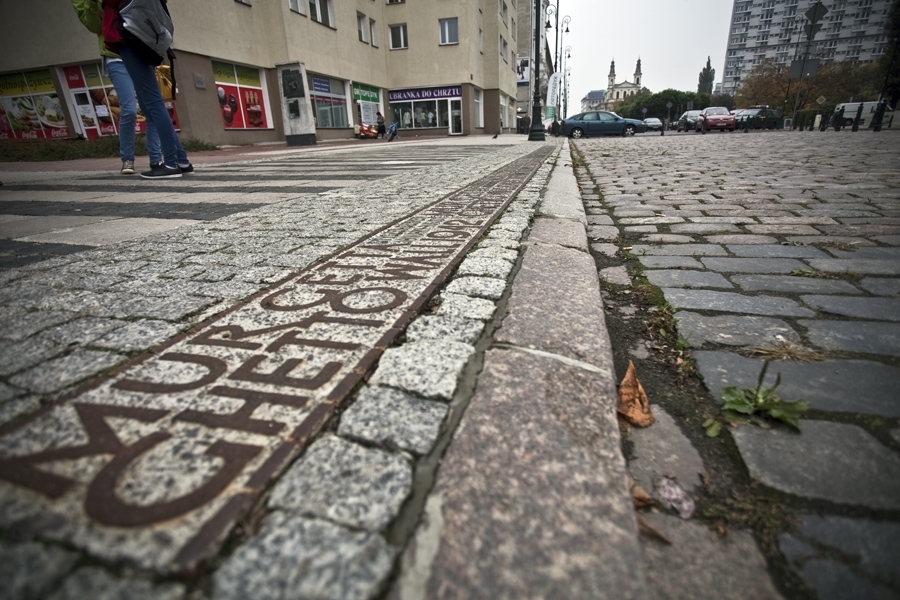 Widok na brukowaną ulicę na Muranowie. Zbliżenie na metalową opaskę, wmurowaną w bruk, która określa, jak przebiegały mury getta warszawskiego