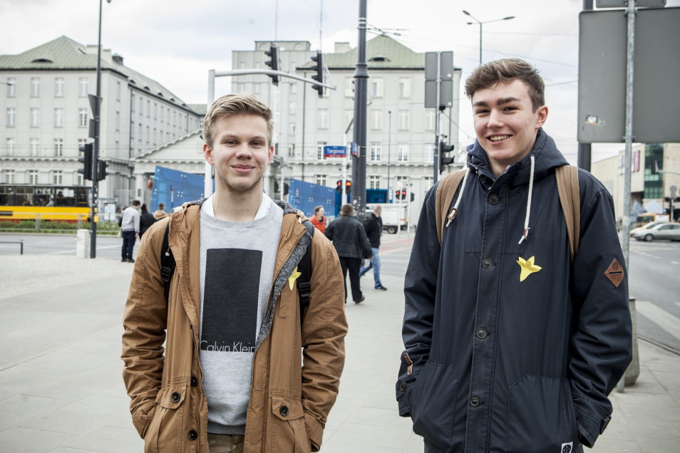 Dwóch chłopaków uśmiechniętych na ulicy Warszawy, niedaleko metra z przylepionymi żonkilami