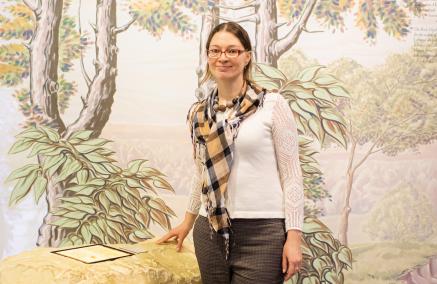 Alla Marchenko - szczupła brunetka w okularach, ubrana jest w białą bluzkę, dżinsy i szalik w kratkę. Stoi na tle ściany z namalowanymi drzewami. 