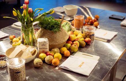 Na blaszanym stole leżą owoce, warzywa i serwetki z napisem POLIN. Stoją też na nim przyprawy w słoiczkach, mąka, naczynie z ziołami, wazon z tulipanami.
