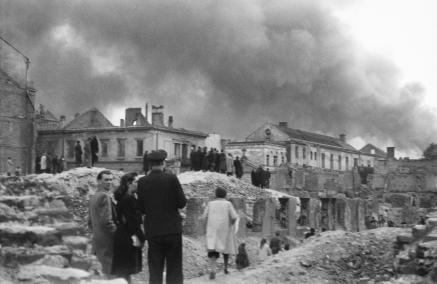 Kilka osób stoi przy zniszczonych zabudowaniach szpitala św. Ducha. W tle dym płonącego getta.