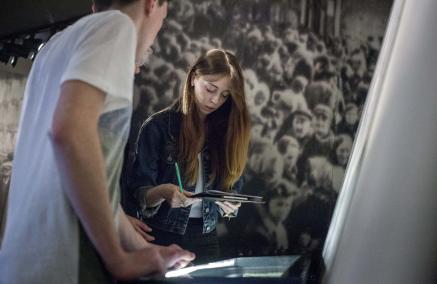 Dziewczyna i chłopak stoją przy gablocie na wystawie stałej w Muzeum POLIN. Dziewczyna trzyma notes i długopis.
