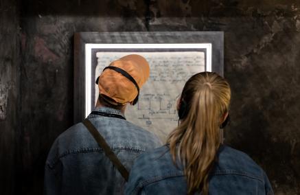 Chłopak i dziewczyna czytają fragment dziennika na wystawie "Wokół nas morze ognia". Stoją tyłem, ich twarze zwrócone w stronę ściany.