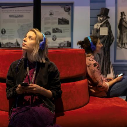Dwie kobiety siedzą na kanapie w jednej z galerii na wystawie stałej w POLIN. Mają słuchawki na uszach i słuchają nagrań z audioprzewodnika.