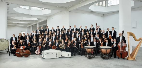 Na zdjęciu widać muzyków Orkiestry Sinfonia Varsovia w strojach koncertowych, wraz z instrumentami. Muzycy znajdują się w dużej jakby fabrycznej przestrzeni, z wieloma ogromnymi świetlikami w suficie