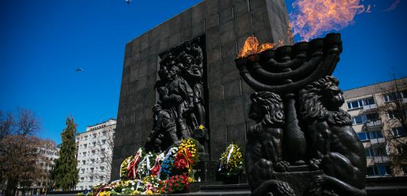 Pomnik Bohaterów Getta, płonie ogień na zniczu w kształcie menory, u stóp pomnika leżą kwiaty
