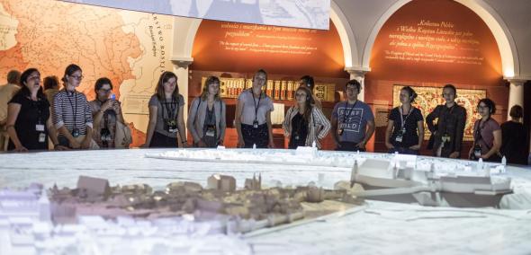 Grupa młodzieży stoi przed ogromną, podświetloną makietą średniowiecznego Krakowa. W tle ściany wystawy "1000 lat historii Żydów polskich" Muzeum POLIN