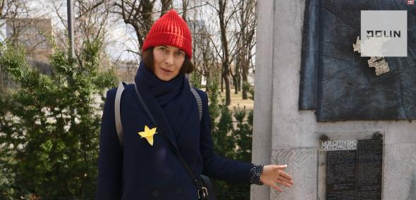 Na zdjęciu kobieta (Beatat Chomątowska) z przypiętym do płaszcza żółtym papierowym żonkilem - symbolem prowadzonej przez Muzeum POLIN akci Żonkile - stoi przed jednym z pomników poświęconych upamiętnieniu getta warszawskiego