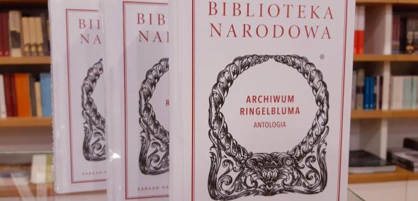 Okładka książki "Archiwum Ringelbuma. Antologia" w wydaniu Biblioteki Narodowej seria "Ossolineu". Ustawiona pionowo na stole. W tle regał z innymi książkami