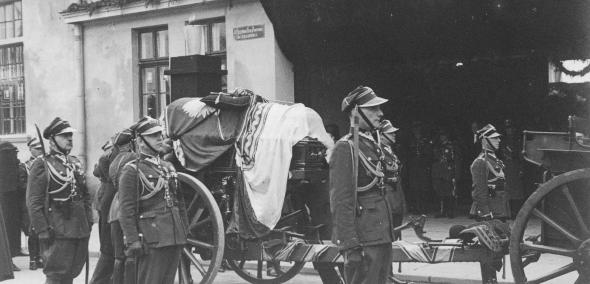 Zdjęcie archiwalne - żołnierze w mundurach galowych stoją na baczność wokół lawety. Na lawecie trumna z ciałem Józefa Piłsudskiego przykryta flagą Polski. Na trumnie leży czapka Piłsudskiego i jego szabla.