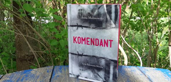 Okładka książki: Anna Malinowska "Komendant. Życie Salomona Morela". Książka stoi na stoliku w ogrodzie, wśród zieleni