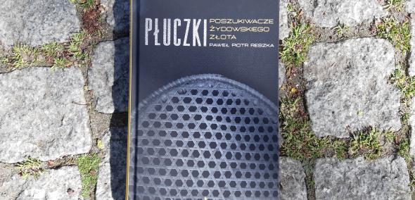 Okładka książki Paweł Piotr Reszka "Płuczki. Poszukiwacze żydowskiego złota". Książka leży na bruku ulicy