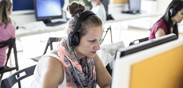 Na zdjęciu kobieta w słuchawkach na uszach. Siedzi przed monitorem komputera.
