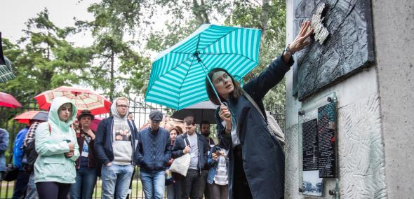 Na zdjęciu kobieta (przewodniczka) pokazuje element architektury i omawia go grupie zwiedzającej młodzieży. Deszczowy dzień, ludzie w kurtkach przeciwdeszczowych, pod parasolami