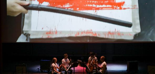 Scena, kwartet smyczkowy, nad muzykami ekran, na którym wyświetlane jest wideo" policyjną gumową pałką czyjaś ręka uderza w białą płachtę, powstaje na środku krwawy czerwony pas - płachta zaczyna wyglądać jak flaga Białorusi