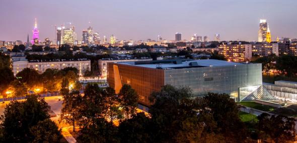 Budynek Muzeum widziany z góry, z budynków przy ulicy Edelmana. Jest wieczór, widać rozświetloną panoramę śródmieścia Warszawy