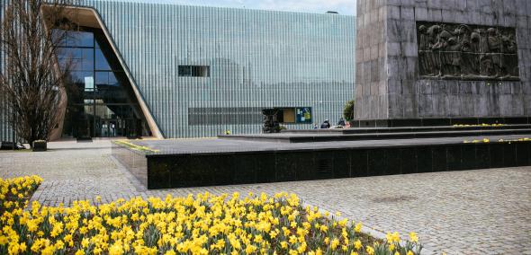 Z prawej strony w głebi budynek muzeum, widać wejście. Bliżej, po prawej, Pomnik Bohaterów Getta. Na pierwszym planie po lewej widać usiany żółtymi żonkilami trawnik.