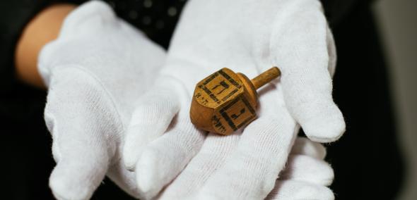 Osoba w białych rękawiczkach trzyma czworoboczny bączek chanukowy (drejdel) wykonany z drewna, z literą hebrajską na każdym boku.