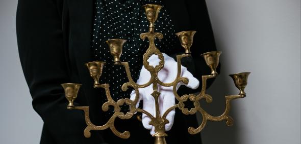 Osoba w białych rękawiczkach trzyma stojący świecznik synagogalny siedmioramienny w formie trójkąta.