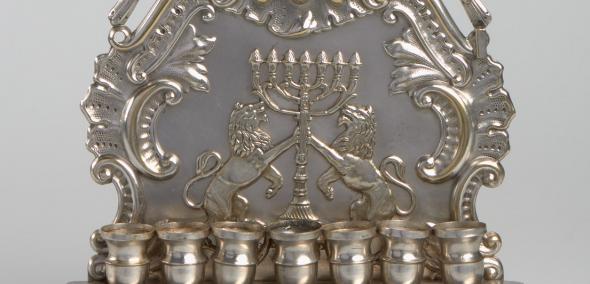 Chanukija składająca się z podstawy o czterech ażurowych nóżkach, na której umieszczone zostało 8 nasadek na świece. Za nasadkami umieszczony został  pionowy zaplecek ozdobiony płaskorzeźbami prezentującymi odwołujące się do symboliki żydowskiej przedstawienia. 