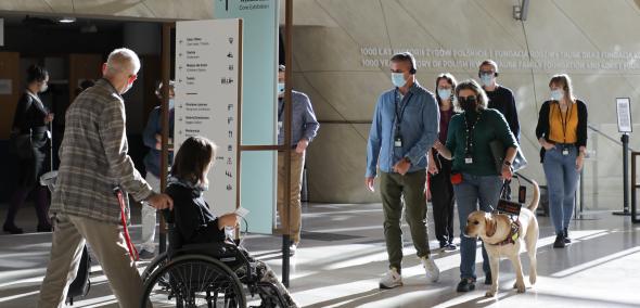 Zwiedzający z niepełnosprawnościami – niewidomi, osoba na wózku, z psem przewodnikiem –  w holu Muzeum POLIN.