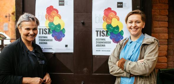 Dwie kobiety stoją przy drzwiach z plakatami Tisz festiwalu 2020. Obie uśmiechają się.
