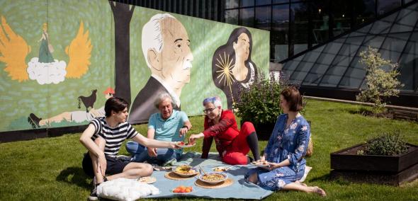 Cztery osoby piknikują na łące przy Muzeum POLIN. Na kocu leżą miski z różnymi przysmakami, np. hummusem.