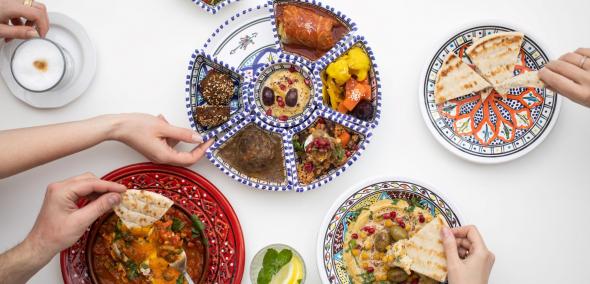 Na stole znajdują się talerze z mezze izraelskimi - pitą, hummusem, falafelami, tahini - i miska z szakszuką. Częstuje się nimi kilka osób - nad talerzami wyciągnięte dłonie.