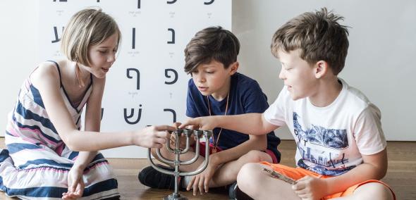 Troje dzieci siedzi na podłodze. Dotykają świecznika. Za nimi tablica z literami alfabetu hebrajskiego.
