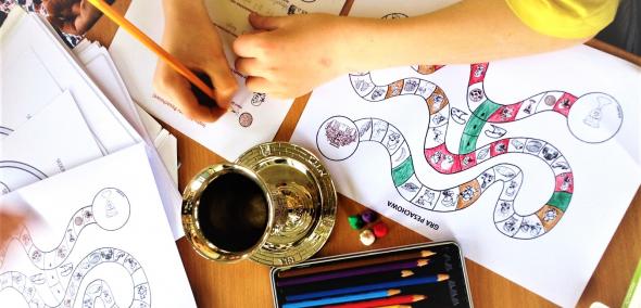 Kolorowanki, kielich i kredki leżą na stole. Dziecko maluje jedną z kolorowanek.
