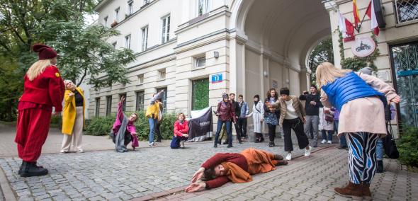 Grupa osób stoi w półkole przy jednej z kamienic na warszawskim Muranowie. Pomiędzy tymi osobami na chodniku leży kobieta.