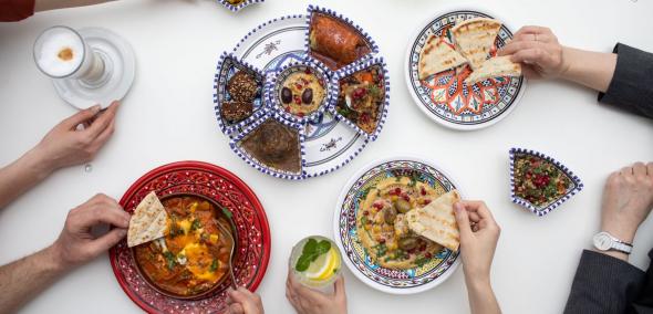 Na stole w miskach stoją potrawy: pita, szakszuka, humus, gołąbki, sałatka izraelska. Kilka osób degustuje potrawy.