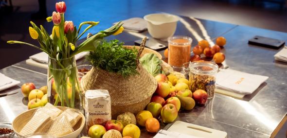Na blaszanym stole leżą owoce, warzywa i serwetki z napisem POLIN. Stoją też na nim przyprawy w słoiczkach, mąka, naczynie z ziołami, wazon z tulipanami.