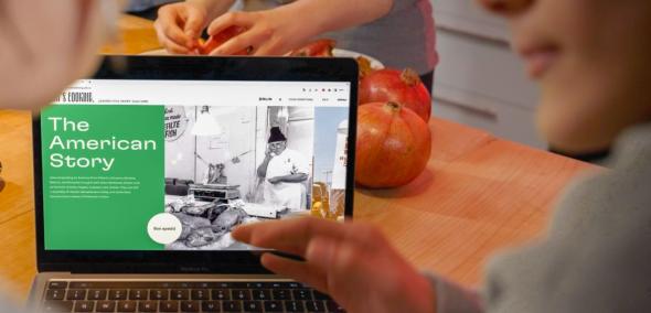 Zza pleców dwóch osób widać ekran laptopa, który stoi na blacie stołu. Na ekranie wyświetlona strona www "Od kuchni". Widać napis po angielsku: The American Story