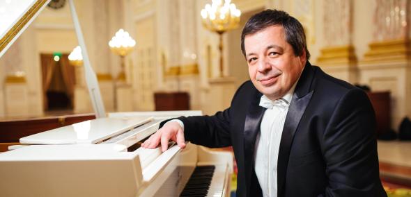 Aleksey Botvinov siedzi przy białym fortepianie w sali ze świecznikami.