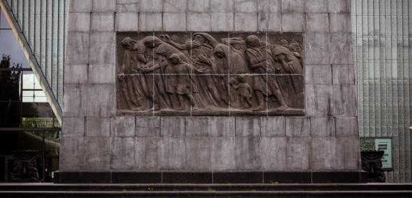 Druga strona pomnika Bohaterów Getta Natana Rapoporta - płaskorzeźba przedstawiająca ludność cywilną w pochodzie na Zagładę. W tle budynek Muzeum POLIN.