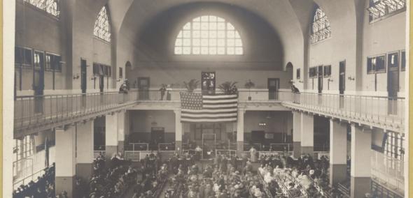 Grupa ludzi siedzi w sali wykładowej. Na górze wisi flaga USA. Zdjęcie archiwalne.