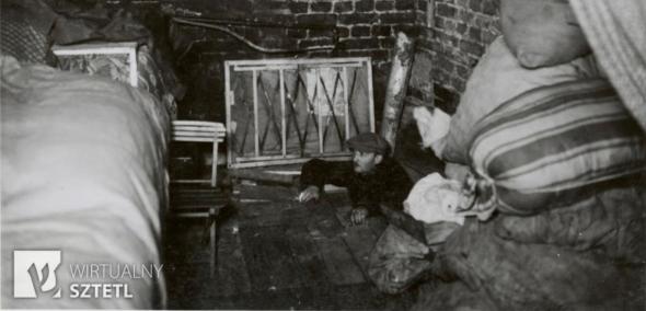 Kryjówka w getcie warszawskim w czasie powstania 1943 roku. Wewnątrz łóżko, worki, naczynia. Do środka przez klapę wchodzi jakiś mężczyzna w kaszkiecie.